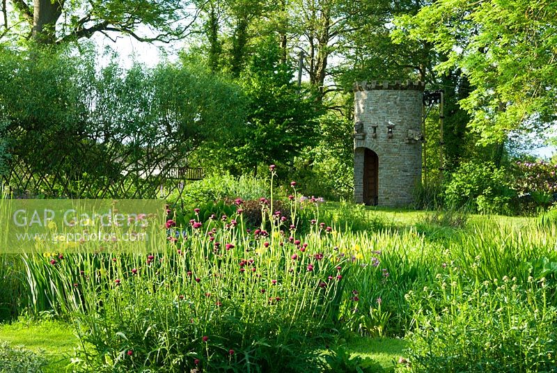 Stone dovecot that operates as a spectacular water feature as well. A simple belt and trough system scoops water up from the mill leat into a tank which, when full spouts throught the open mouth of a gargoyle back into the mill pond. Clump of deep red Cirsium rivulare 'Atropurpureum' in foreground - Westonbury Mill
 