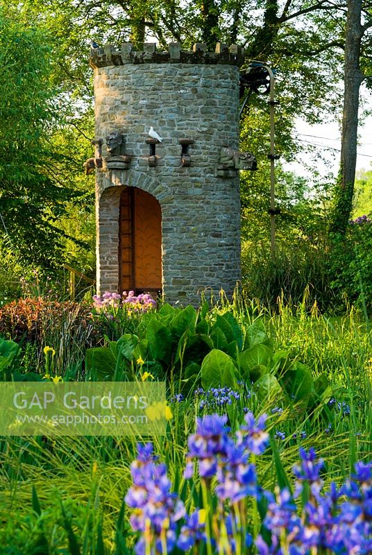 Stone dovecot that operates as a spectacular water feature with Iris sibirica in foreground. A simple belt and trough system scoops water up from the mill leat into a tank which, when full spouts throught the open mouth of a gargoyle back into the mill pond - Westonbury Mill