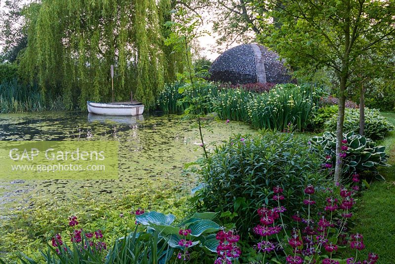 Lake edged with Hostas and Primulas contains small rowing boat below a Salix - Weeping willow tree, with dome shelter made of glass bottles beyond - Westonbury Mill