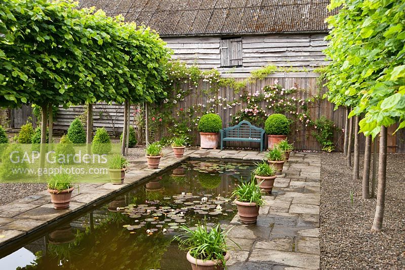 The Canal, framed by pleached limes and pots of agapanthus, with seat at end surrounded by pink Rosa 'Albertine' on right and Rosa 'Paul's Himalayan Musk' on the left. Ashley Farm, Stansbatch, Herefordshire, UK