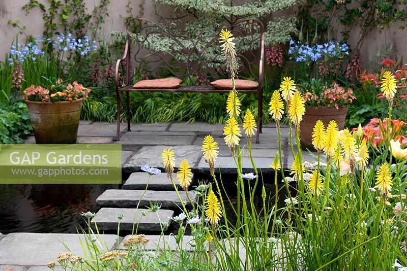 Beds and pots with mixed herbaceous planting including Kniphofia 'Brimstone' and central pool with stepping stones leading to intimate seating area in the 'Summer Fairytale' garden. RHS Tatton Flower Show 2013