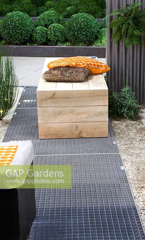 Metal floorgrate and wooden box as seat with orange and brown pillows. Buxus balls in border