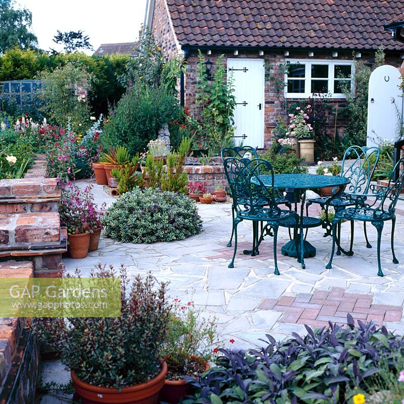 Patio of crazy paving and bricks with metal table and chairs and container planting throughout