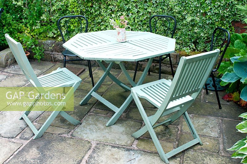 Small slatted wooden table and chairs on stone patio and sheltered by wall coveed with Hedera