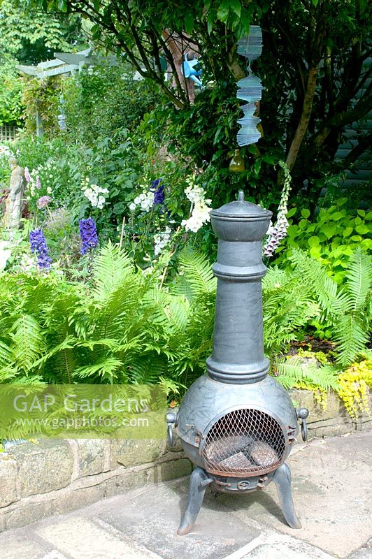 Chiminea on stone patio backed by raised bed with fern Digitalis and Delphinium in cottage garden