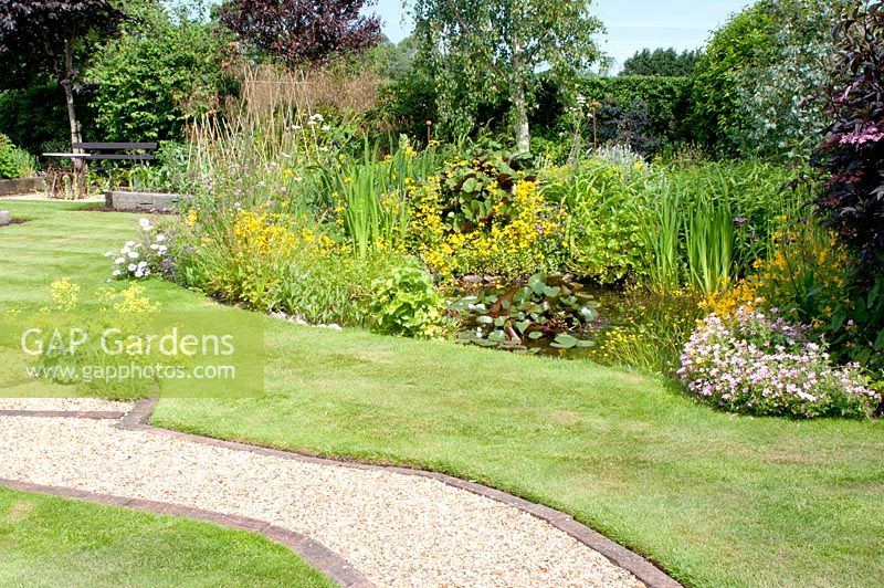 Gravel path edged by block paving and natural garden pond with Nymphaea Iris Caltha Mimulus - Monkey flower and Geranium in mature cottage garden 