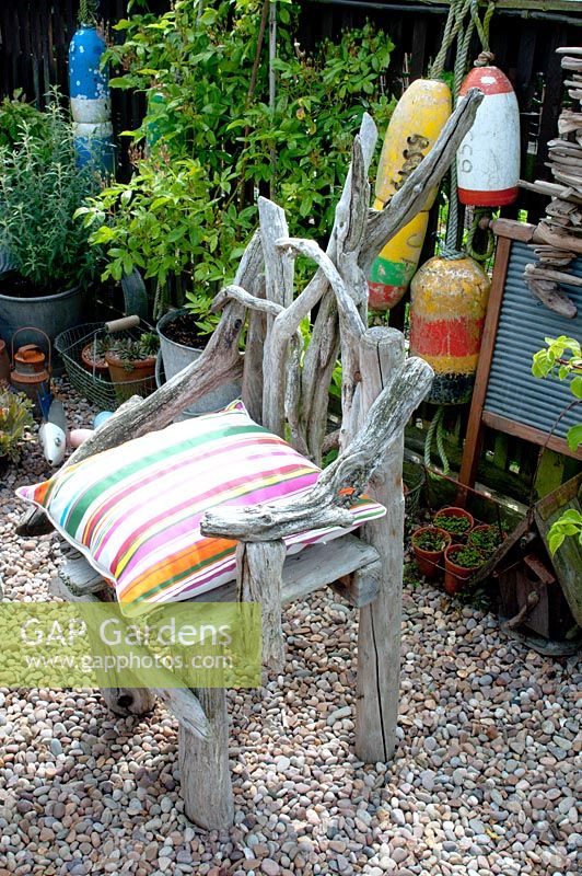 Chair made of driftwood on gravel surface in corner of cottage garden surrounded by vintage collection of galvanised planted containers and fishing floats against wooden fence
