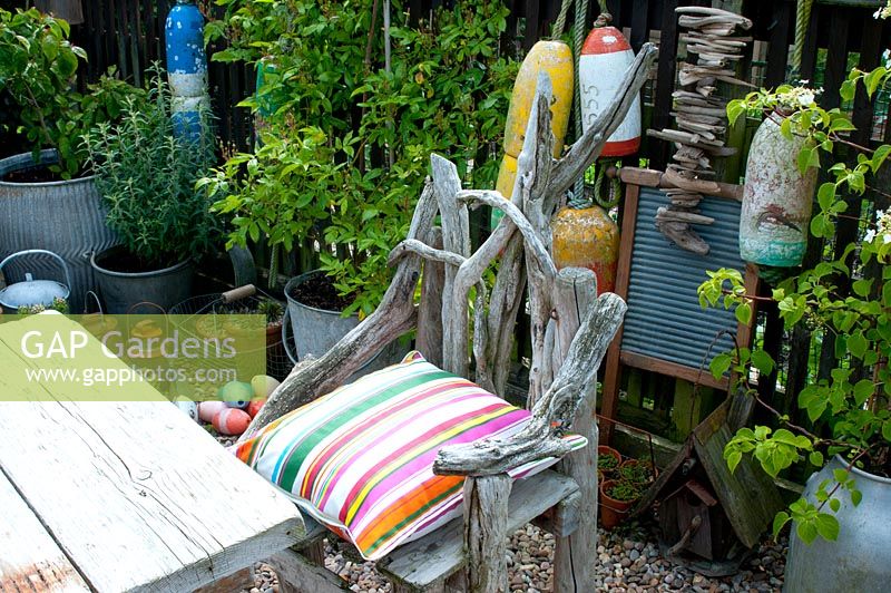 Chair made of drift wood - corner of cottage garden surrounded by vintage collection of galvanised planted containers and fishing floats against wooden fence  