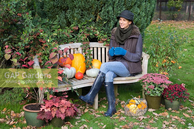 Woman sat on garden bench with autumnal display of pumpkins. 'Crown Prince', 'Mammoth', 'Uchiki Kuri' and basket of Gourds.