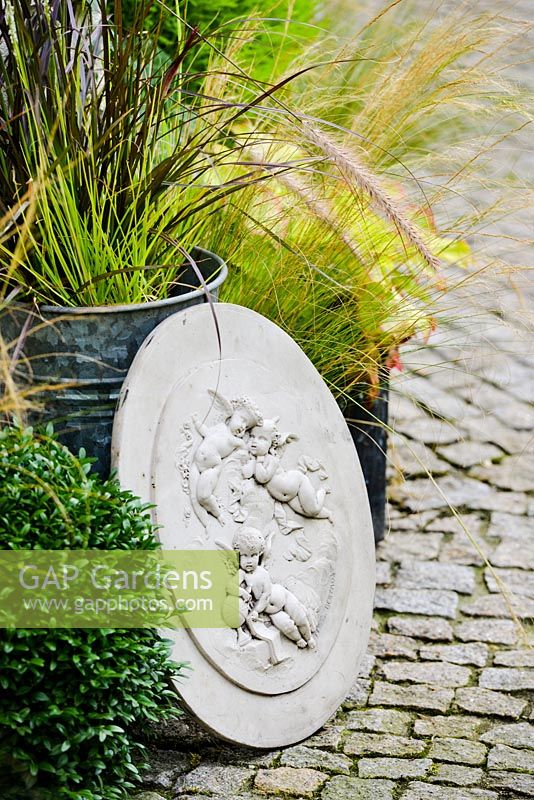 Engraved concrete plaques with angels figures leaning against flower pots with grasses on granite setts path.