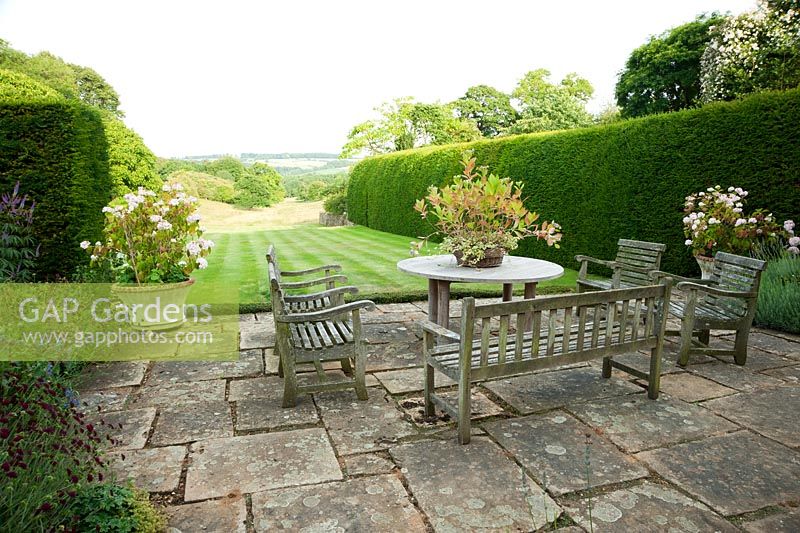 Terrace framed with hydrangeas in pots, with lawn below edged by tall yew hedges, and meadow in the valley beyond. 