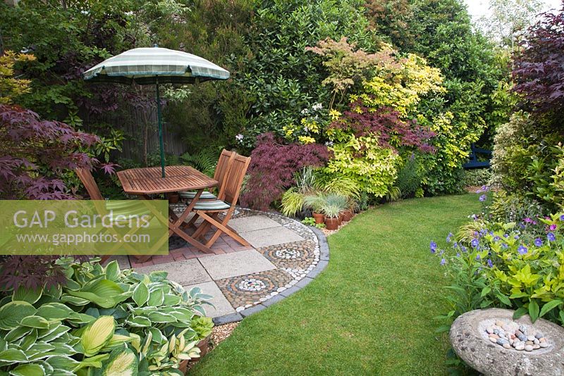 Seating area with mosaic effect stonework on patio and mixed colourful foliage of Acer, Hosta and shrubs in summer 