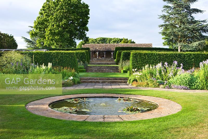Circular pond in the lawn with view through hedges to the cotswold stone loggia