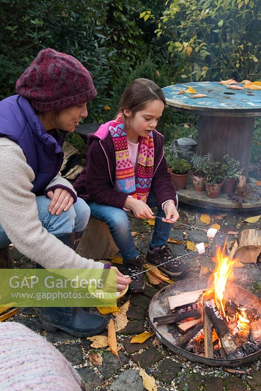 Mother and daughter roasting marshmallows over a firepit in an autumnal back garden.