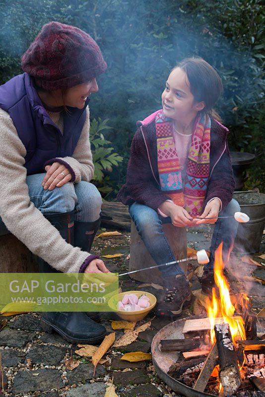 Mother and daughter roasting marshmallows over a firepit in an autumnal back garden.