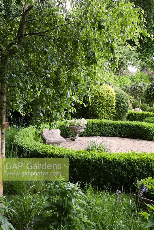 Designed topiary structures in formal country garden including horseshoe low Buxus hedge around circular patio with stone seat