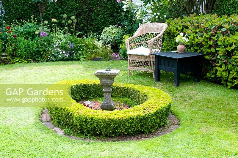 Wicker chair and table on lawn in cottage garden with low circular clipped Buxus hedge surrounding stone sundial
