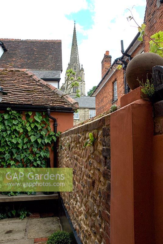 Small town garden with Ivy clad wall, wooden bench and box ball beneath a flint and brick wall. York stone paving and a Church spire in background