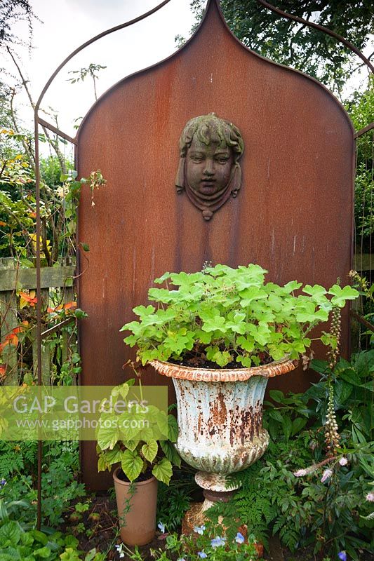 Metal feature in the Parterre Garden with cherub's head and urn with pelargonium. Whimble Garden and Nursery, Kinnerton, Powys, Wales