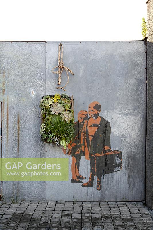 Planting display in vertical container hanging on wall with painting of children