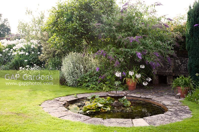 Decorative stone circular pond in summer with fountain and planting including Phlox, Phalaris arundinacea, Buddleia and Agapanthus