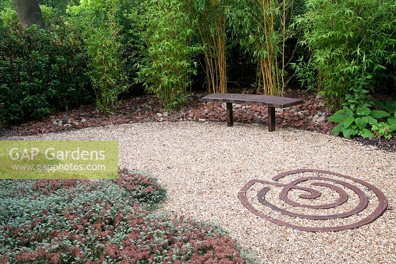 Decorative metal feature on ground in gravel in Rust Garden with metal bench. Plants include Phyllostachys aureosulcata 'Spectabilis' and Acaena microphylla 