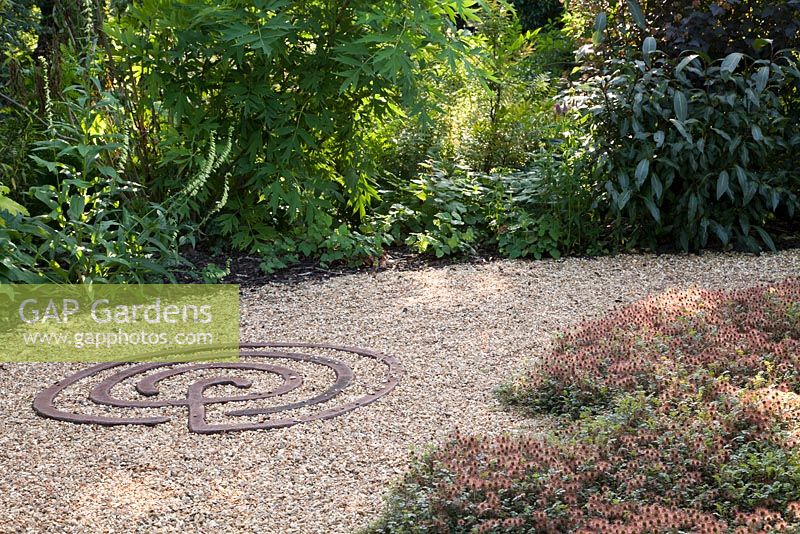 Decorative metal feature on ground in gravel in Rust Garden with Acaena microphylla