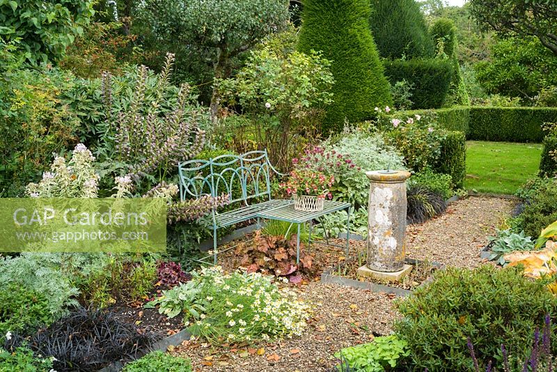 Formal garden in late summer with blue painted wrought iron bench, old sundial, gravel paths, roses, herbaceous perennials and view to lawns with box hedging and Yew topiary.