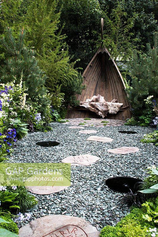The Viking Cruises Norse Garden.  View of path with Viking rune stepping stones, small fountains and slates stones leading to Viking Longship Garden Sculpture with seat and leather surrounded by Anchusa 'Lodden Royalist', Digitalis 'Dalmation White' and Pines trees. 