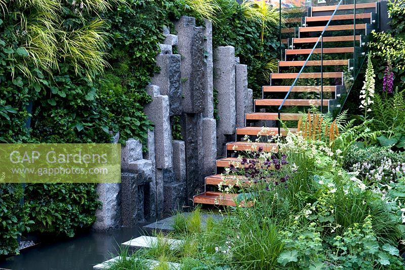 Green living wall with hart's tongue ferns surrounds lengths of rough-hewn granite stone uprights down which dribbles water. A wooden open-work staircase leads to upper mezzanine. Loosely planted border of shade-loving perennials. The Mind's Eye garden for the RNIB, gold medal winner. RHS Chelsea Flower Show 2014. 
