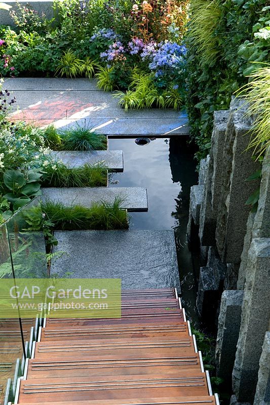 View from upper mezzanine down staircase to granite stone flooring into which is set shallow black water pool. Shade-loving plants in geometric beds. The Mind's Eye garden for the RNIB, gold medal winner. RHS Chelsea Flower Show 2014. 