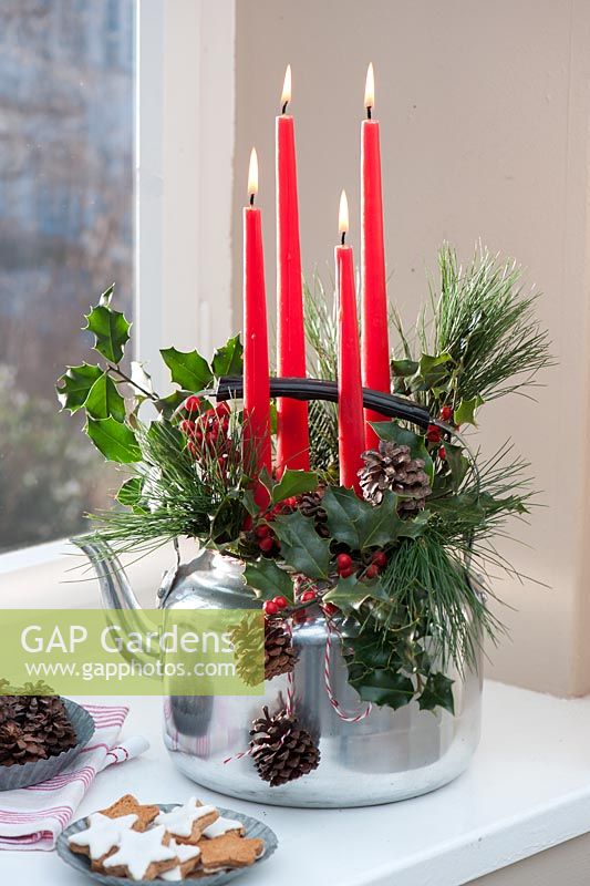 Christmas candles decorated with Holly sprigs and pine cones in metal teapot
