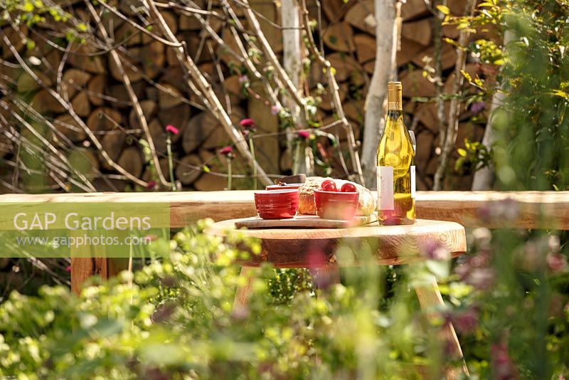 Rustic wooden bench and table with food and wine with split log wall beyond, 'Shears and Chardonnay', show garden, RHS Malvern Spring Festival 2014
