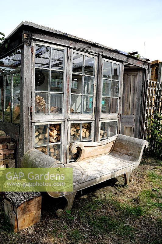 Allotment shed, hut or glasshouse made of stripped wood with wooden chaise lounge in front and logs inside. Golf Course Allotments, London Borough of Haringey.