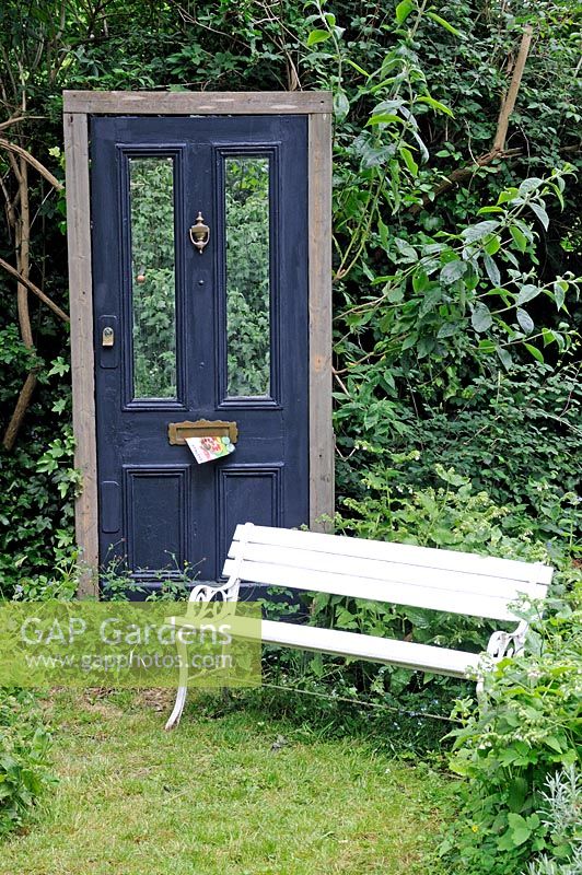 Victorian front door used as Trompe l'oeil in a garden in Hackney, with junk mail in letterbox and white painted bench to fore.