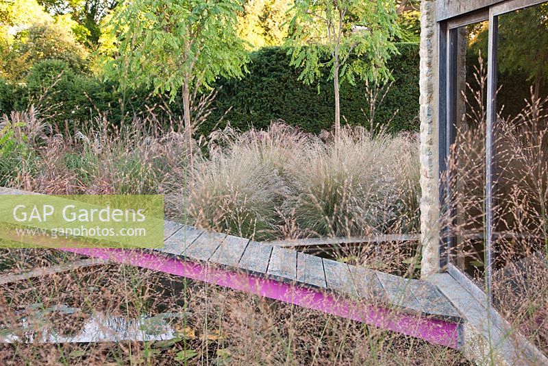Decorative pink steel and wooden bridge over pond to summerhouse through Molinia 'Transparent' - Farrs
