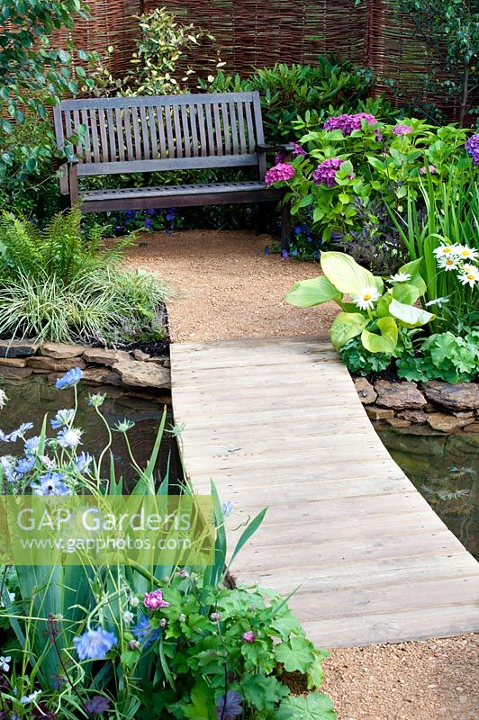 Wooden bridge over pool leading to seating area with bench and colourful beds in the 'Ripples on The Water' garden designed by Muddy Boots - Southport Flower Show 2013