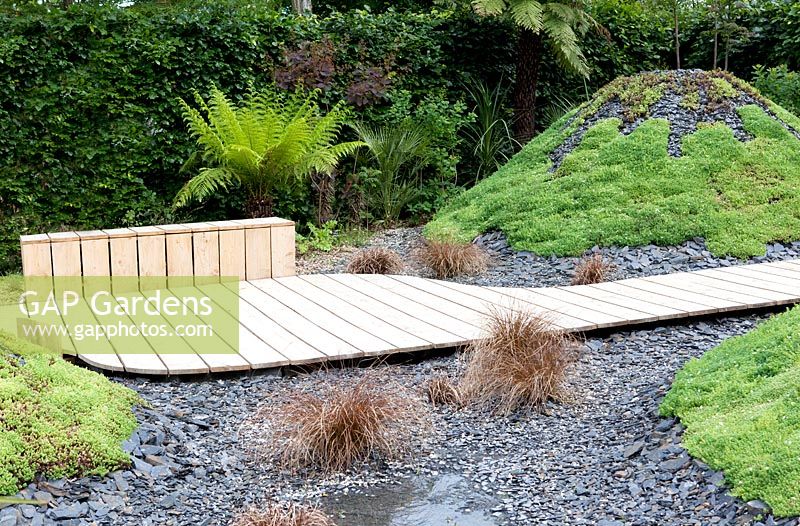Title: Pour L Amour de Tongariro. Garden with wooden path passing several 'volcanos' planted with sedums and covered with broken black slates.