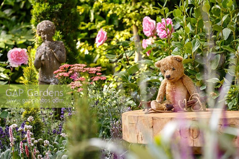 Old fashioned teddy bear and skipping rope on wooden bench amongst 1920s period planting and statue - The NSPCC Legacy Garden, RHS Hampton Court Flower Show 2014