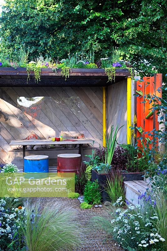 A Space To Connect and Grow. Wooden shelter area with living green roof, recycled oil drum stools, timber table, bamboo water feature, planting in water trough and recycled timber seat.