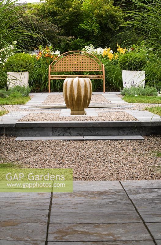 Raised gravel bed with a gourd-shaped water fountain and brown metal lattice garden bench with white Hydrangea paniculata ‘Quick Fire' and Daylily (Hemerocallis). Box (Buxus 'Green Velvet') shrubs in planters - Il Etait Une Fois garden, Monteregie, Quebec, Canada.