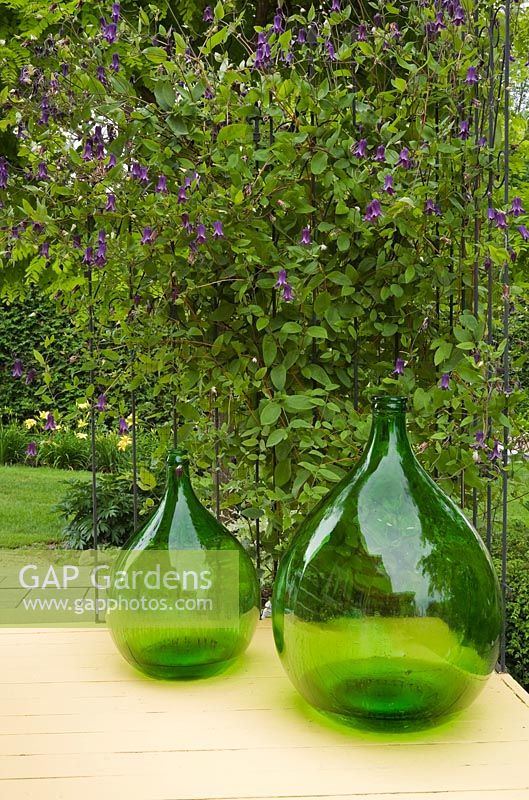 Two large green glass jugs next to a metal trellis with mauve Clematis flowers on a yellow painted wooden plank back porch - Il Etait Une Fois, Quebec, Canada.