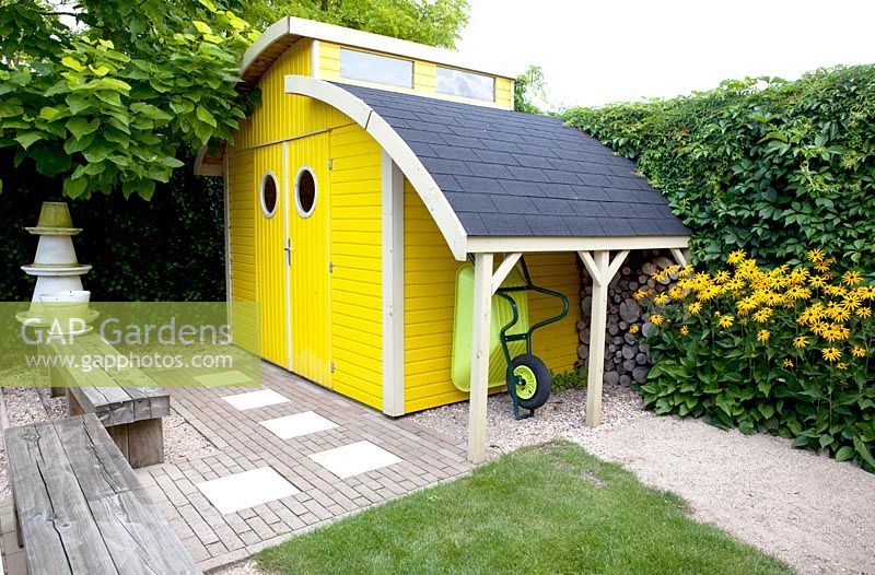 Bright yellow shed with roundshaped windows. Rudbeckia fulgida Goldsturm in the border. Moveable wooden benches on rails.