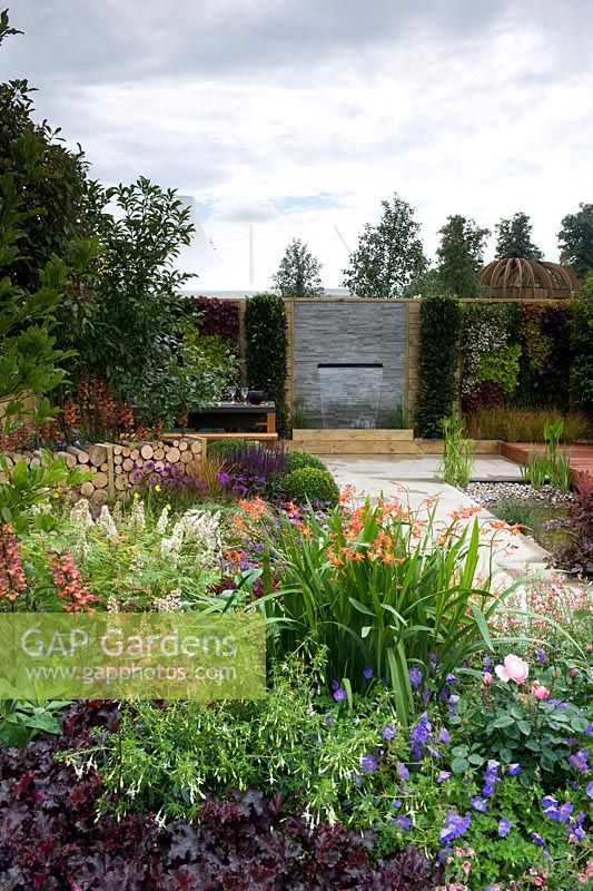 A Hampton Garden - View into garden over border of mixed perennials including heuchera, crocosmia, digitalis and annual viola and fuchsia. A high boundary with water feature and living wall is behind. 