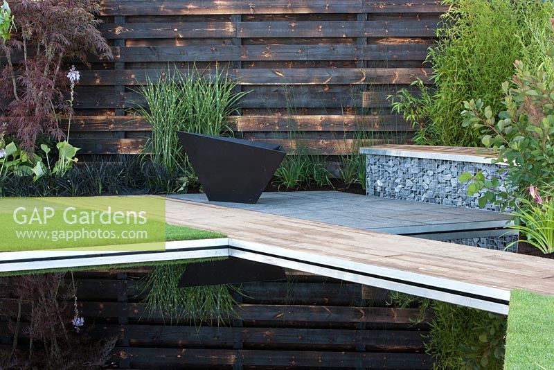 Elemental - reflective dark pool pond with charred burned redwood board wall basalt cobbles in cage with timber seating and path firepit - planting of grasses including Miscanthus sinensis 'Zebrius' zebra grass and bamboo Phyllostachys aurea 