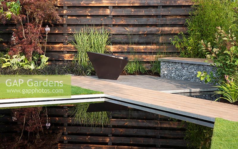 Elemental - reflective dark pool with charred burned redwood board wall basalt cobbles in cage with timber seating and path firepit - planting of grasses including Miscanthus sinensis 'Zebrius' zebra grass and bamboo Phyllostachys aurea