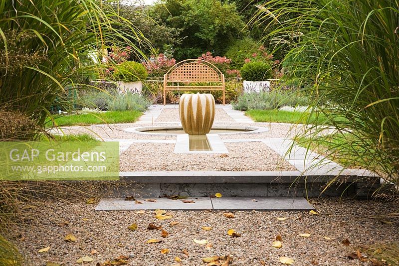 Raised gravel bed with gourd-shaped water fountain and brown metal lattice garden bench with pink hydrangea paniculata 'Quick Fire' framed by tall Miscanthus sinensis 'Berlin' ornamental grasses in backyard garden in autumn. Il Etait Une Fois garden, Monteregie, Quebec, Canada. 