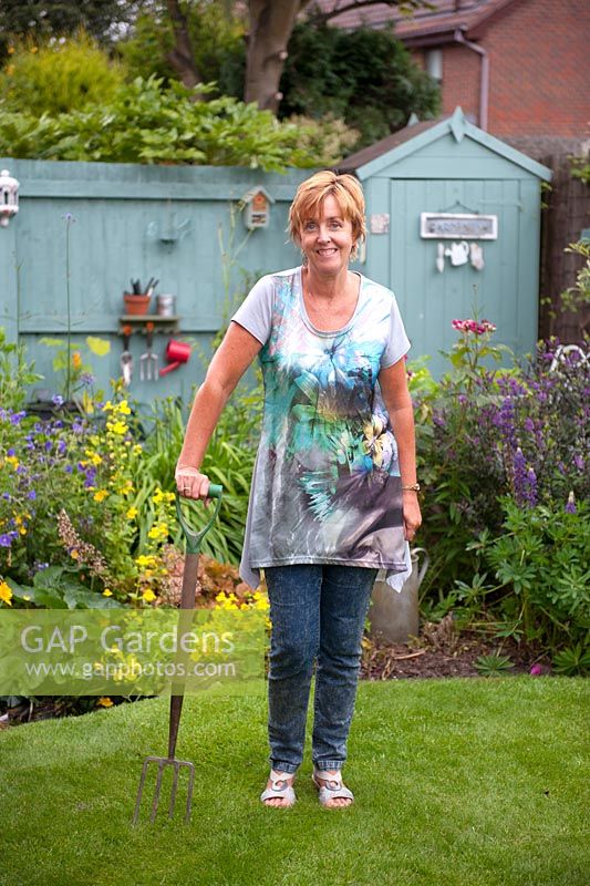 Garden designer, Angie Barker rests on garden fork in front of cottage garden border with Calthra palustris, Lupinus 'Gallery Blue' and small wild life pond