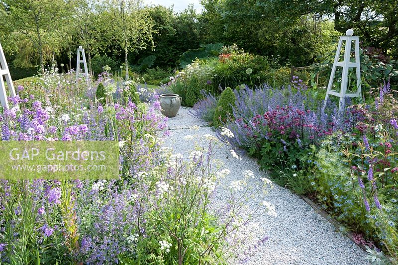 Gravel pathway thorugh flower garden planted with a mix of flowering herbaceous perennials with white wooden obelisks to give year round structure.