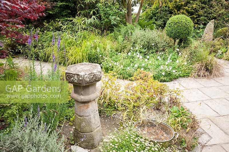 A courtyard garden largely planted with for foliage features clipped box, grasses, euphorbias, corokia and acers around standing stones, architectural fragments and old galvanised tubs used as ponds, planters or mini water gardens. 